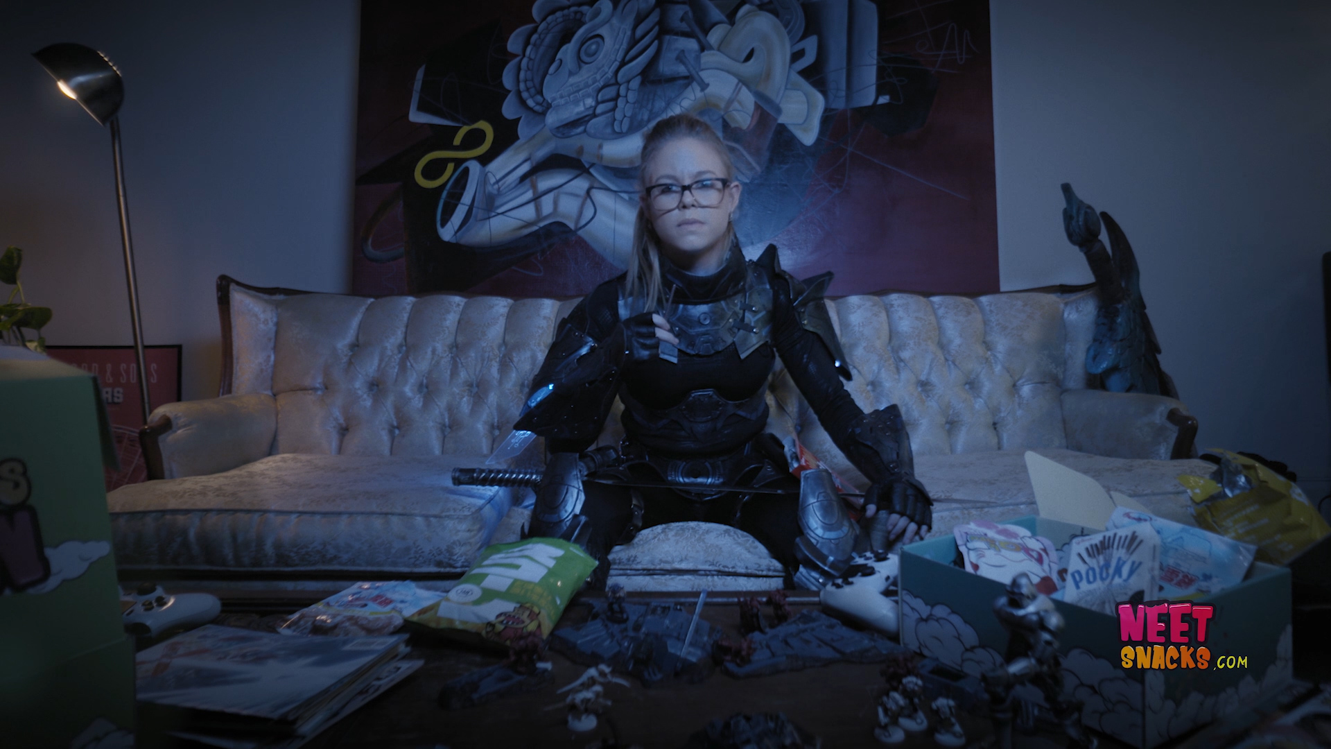 A young girl in glasses in science fiction armor sits on the couch of a dorm room. A board game and snacks is on the coffee table in front of her.